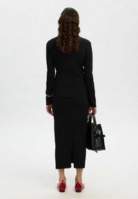 Top fitted a coste nera e gonna midi con spacco posteriore, abbinata a scarpe rosse senza lacci. Tiene una piccola borsa a tracolla nera con manico circolare.