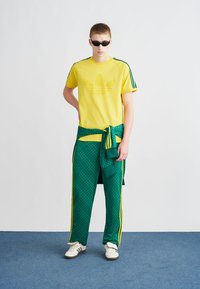 Geltonos spalvos trumpomis rankovėmis marškinėliai su centrinėmis logotipu; žali raštuoti sportinės kelnės su geltonomis juostelėmis; dėvimi su akiniais nuo saulės ir baltais sportbačiais.