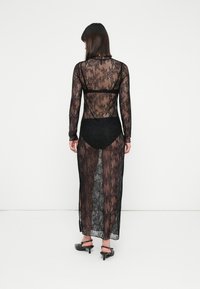 Žena v dlouhých černých průhledných krajkových šatech s dlouhými rukávy a černým spodním prádlem, stojící zády k nám na jednoduchém bílém pozadí.