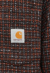 Carhartt-stof i mørkebrun med orange og grå ternet mønster; har et hvidt firkantet logo-patch med et orange ikon og tekst.