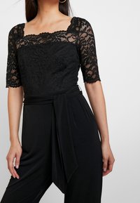 Mono negro con cuerpo de encaje, mangas cortas, escote cuadrado y un cinturón de tela atado a la cintura. Textura suave en general.