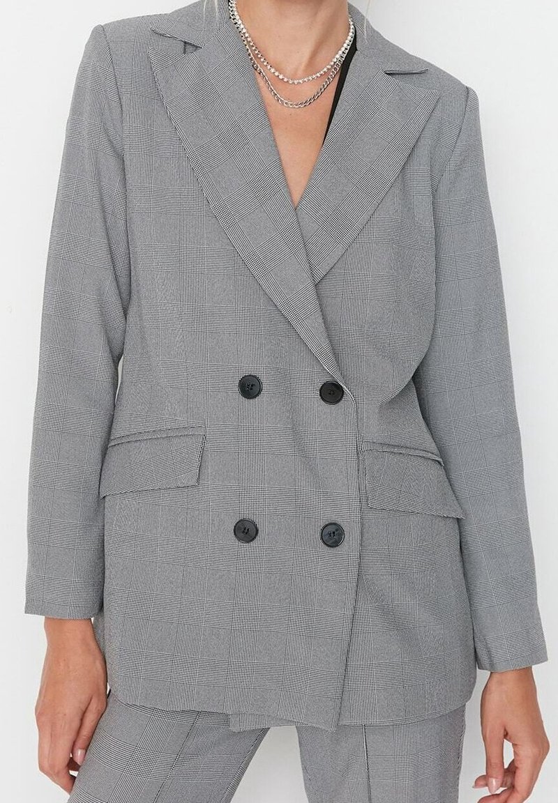 Femme portant un blazer gris à double boutonnage à carreaux et un pantalon assorti, accessoirisée de colliers argentés superposés.