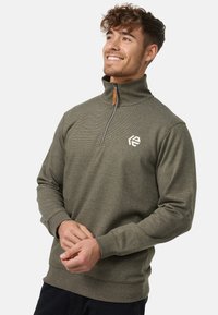 Olivengrøn pullover lavet af tekstureret stof, der har en høj krave, halv lynlås og et lyst logo på brystet. Lange ærmer og ribbet manchetter.