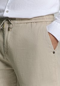 Beige linnenbroek met een elastische tailleband, een trekkoord met metalen uiteinden en zijzakken met knoopaccenten. Textuur van de stof is afgewerkt.