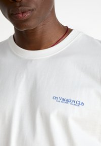 Witte katoenen t-shirt met een blauw bedrukt logo op de linkerkant van de borst dat "On Vacation Club THE SECRET GARDEN" zegt. Korte mouwen.