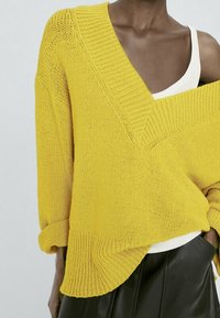 Pull jaune en tricot avec un col en V profond, des poignets et un ourlet côtelés. Porté par-dessus un haut blanc, présentant une coupe asymétrique.