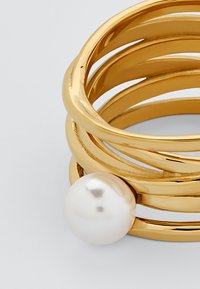 Gullfarget metall armbånd med flere sammenvevde bånd, og har en glatt, blank hvit perle som aksent på den ene siden.
