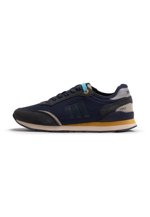 Zapatilla azul marino con detalles en ante negro, raya amarilla en la suela, lazo azul para cordones y "1967" en el panel gris del talón.