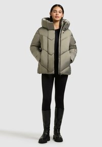 Gepolsterte olivgrüne Jacke mit einer großen Kapuze, Reißverschluss und Zickzackmuster. Getragen über einem schwarzen Oberteil und kombiniert mit schwarzen kniehohen Stiefeln.