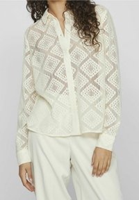 Chemise à boutons en dentelle crème avec motif en losange, manches longues et coupe décontractée. Tissu léger avec détails de design texturés.