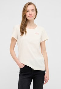 Gai beige t-krekls, kas izgatavots no mīksta kokvilnas, ar īsām piedurknēm, apaļu kakla izgriezumu un nelielu sarkanu "MUSTANG" logotipu kreisajā krūtī.