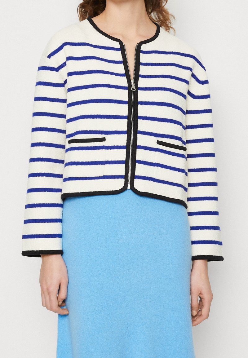 Vit kortad jacka med blå horisontella ränder och svarta detaljer, bärs av en stående person över en ljusblå kjol.