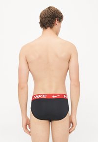 Black swim briefs with a red Nike logo waistband, featuring a smooth texture and form-fitting design, showing the back view.