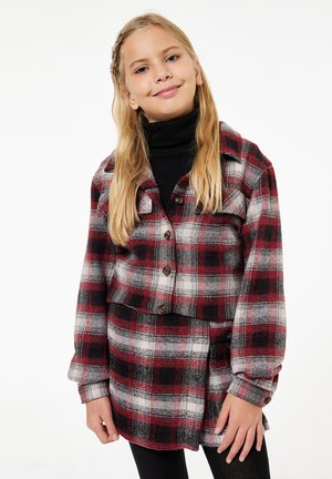 Nuori tyttö, jolla on pitkät vaaleat hiukset, pukeutuneena puna-musta-valkoiseen ruutukuosiseen takkiin ja siihen sopivaan hameeseen mustien leggingsien päällä.