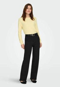 Un pull en tricot jaune à manches longues, associé à un pantalon noir taille haute et une ceinture noire avec une boucle ronde. Des chaussures noires avec un accent en nœud.