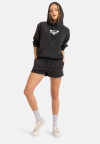 Felpa nera con cappuccio, con lacci e tasca frontale, abbinata a pantaloni corti neri. Il modello indossa calzini grigi e sneakers bianche.