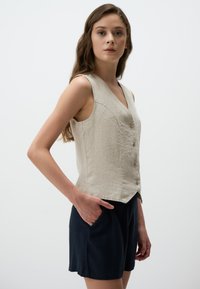 Gilet sans manches en lin beige clair, présentant un décolleté en V profond et cinq boutons. Assorti à un short bleu marine, mettant en valeur une coupe décontractée.
