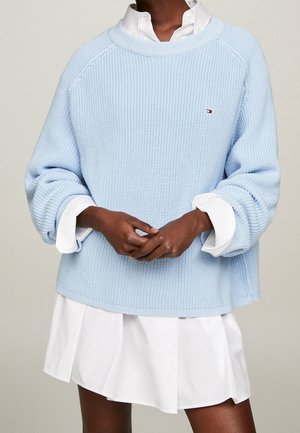 Person trägt hellblauen Strickpullover über einem weißen Hemdblusenkleid und hält die Hände in der Taille verschränkt.
