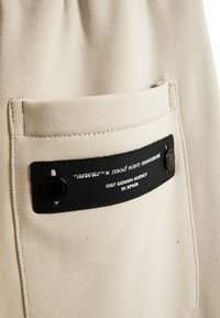 Tela beige con una superficie texturizada, que presenta una etiqueta negra con texto blanco y acentos de metal cerca de un bolsillo lateral.