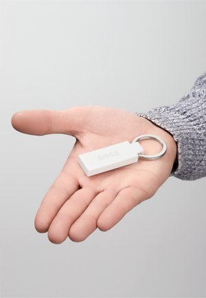 White rectangular keychain with a silver ring, featuring the embossed brand name "BOSS." Held in a hand, against a neutral background.