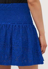 Jupe en dentelle bleue avec une taille froncée, présentant un ourlet à volants superposés et des motifs floraux complexes sur tout le tissu texturé.