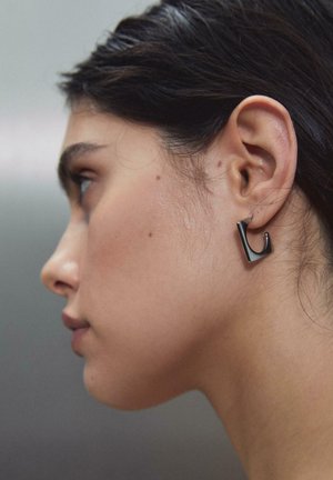 Profil af en kvinde med mørkt hår, der bærer en moderne sort geometrisk ørering på venstre øre mod en sløret neutral baggrund.