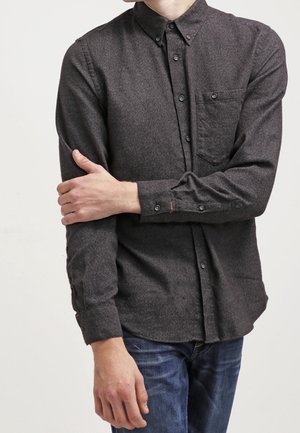 Osoba oblečená v tmavě šedé košili s knoflíky a náprsní kapsou, která si upravuje levý rukáv, doplněná modrými džíny.