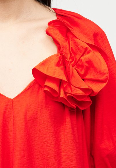 Κόκκινο ένδυμα με μεγάλο, στρωματοποιημένο βολάν στον ώμο και βαθύ V-λαιμόκοψη, φορεμένο από ένα άτομο με ανοιχτόχρωμο δέρμα και σκούρα μαλλιά.