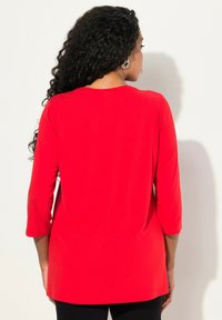 Červený top s dlouhým rukávem a mírně volným střihem, hladkou texturou a zakulaceným výstřihem, s polos dlouhým designem a bez viditelných vzorů.
