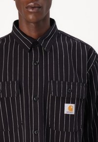 Black and white striped shirt with buttoned collar, featuring two front pockets and a Carhartt logo tag on the left pocket.