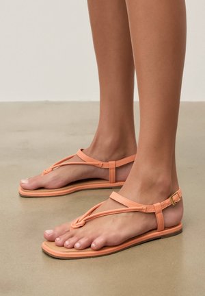 Fødder iført koralfarvede flade sandaler med knyttede remme og ankelspænde på en glat beige gulv.