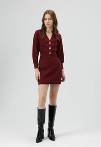 Robe-pull bordeaux à col en V, avec cinq gros boutons et manches longues ; assortie à des bottes noires montantes brillantes à talons.