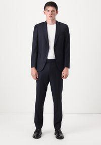 Jeune homme portant un costume à fines rayures foncées avec un t-shirt blanc et des chaussures habillées noires, debout devant un fond blanc uni.