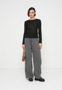 Γυναίκα που φοράει μαύρο μακρυμάνικο τοπ και φαρδιά γκρι τζιν, κρατώντας καφέ τσάντα, στέκεται μπροστά σε απλό λευκό φόντο.