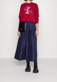 Sudadera roja con gráfico de conejo y texto, combinada con una falda larga de satén azul marino y un bolso de hombro negro. Botas negras de tacón grueso.