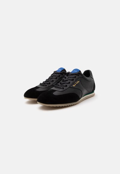 Paire de baskets basses noires avec étiquettes bleues sur la languette, semelles beiges et logo « Paul Smith » sur le côté, sur fond clair.