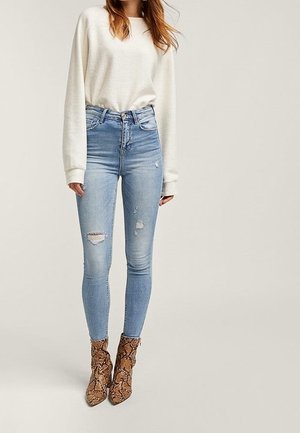 Vrouw draagt een crèmekleurige trui, hoog getailleerde lichtblauwe gescheurde skinny jeans en bruine enkellaarzen met slangenprint, staand tegen een effen achtergrond.