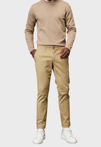 Beżowy sweter z wysokim ściągaczem, zestawiony z jasnymi bawełnianymi chinosami w kolorze khaki i białymi trampkami, z detalem w postaci zapięcia w talii.