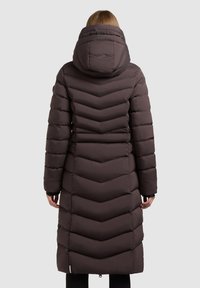 Mørkebrun lang pufferjakke med et chevron quilt mønster, hætte og tætsiddende design. Har ribbede manchetter og en kort lynlås ved kanten.