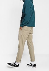 Tyrkysová větrovka s kapucí, síťovanou vložkou a černými akcenty; v kombinaci s béžovými zúženými kalhotami; černé a bílé tenisky; viditelné ohrnuté manžety.