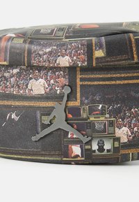 Musta teksturoitu laukku, jossa on koripalloteemaisia grafiikoita, mukaan lukien vintage-televisiot ja hopea Jumpman-logo. Kompakti, suorakulmainen muoto.