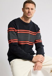 Homme assis portant un pull bleu marine rayé horizontalement de rouge et de blanc, et un pantalon couleur crème, sur un fond uni.