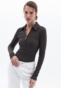 Langärmliges geripptes Oberteil in Dunkelbraun mit einem Reißverschlusskragen. Kombiniert mit hochtaillierten weißen Jeans und einem dezenten silbernen Knopfdetail.