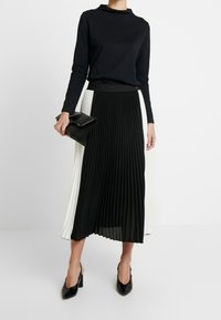 Zwarte langarmtop gecombineerd met een geplooide rok met afwisselend zwarte en witte panelen. Houdt een zwarte clutch vast en draagt zwarte hakschoenen.