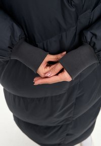 Puffer jakna u tamnoplavoj boji s naglašenim rukavima i sivim rebrastim završecima. Ima teksturirani, quiltani dizajn. Ruke su spojene iza leđa.