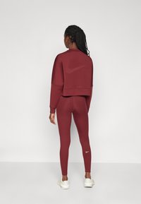 Costum sportiv burgund care include un hanorac scurt cu un logo tonal și colanți asortați, realizat dintr-un material neted și elastic.