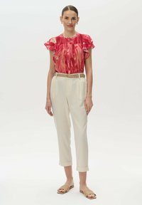 Rote, gemusterte Bluse mit Rüschensäumen, kombiniert mit hellbeigen, umgeschlagenen Hosen. Ein beiger Gürtel und flache Sandalen runden das Outfit ab.