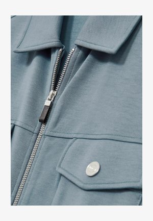Light blue zip-up jacket with a collar, featuring silver hardware, a textured fabric, and two front pockets with snap closures.