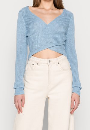 Woman wearing a light blue knitted crossover crop top with long sleeves and high-waisted off-white wide-leg pants.