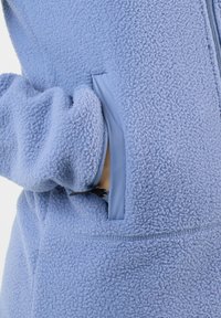 Giacca in pile azzurro chiaro con superficie texture, tasca con zip e polsini elasticizzati. Dettagli includono un tessuto interno liscio.